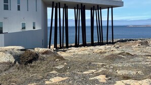 A modern building with a flat roof supported by vertical pillars, located on a rocky shoreline with a view of the sea.
