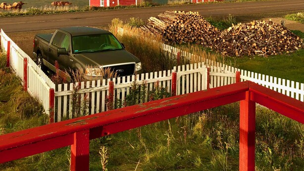 A rural landscape with a truck parked near a white picket fence, a red barn, and a rainbow in the sky.