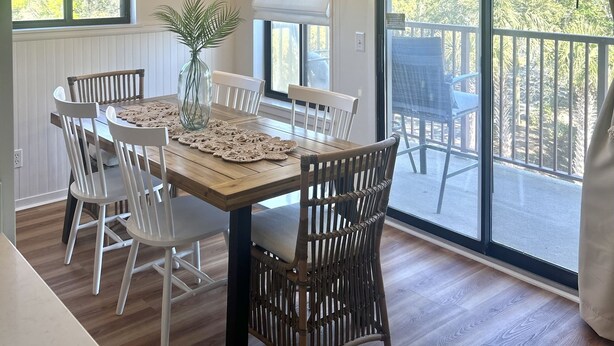 Un coin repas avec une table en bois, des chaises blanches, un fauteuil en osier, un lustre et une vue sur un balcon aménagé avec du mobilier d’extérieur.