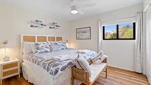 Une chambre à coucher comprenant un lit, des tables de chevet, un ventilateur de plafond, une fenêtre avec des rideaux et des œuvres d’art murales.