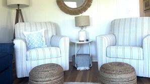 Un salon avec deux fauteuils à rayures, un miroir rond, une petite table d’appoint avec une lampe et deux poufs en osier posés sur un tapis à motifs.