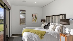 Quarto com cama, duas luminárias de cabeceira, mesa de apoio, janela com persianas e porta para área externa.