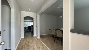 A hallway leading to a dining area with a table and chairs, a door on the left, and a ceiling light.