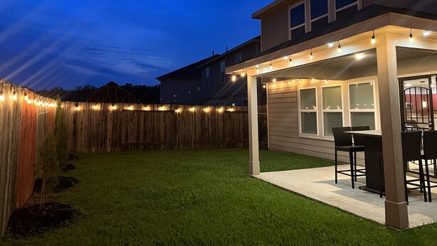 A backyard patio with a covered seating area, a wooden fence, and string lights.