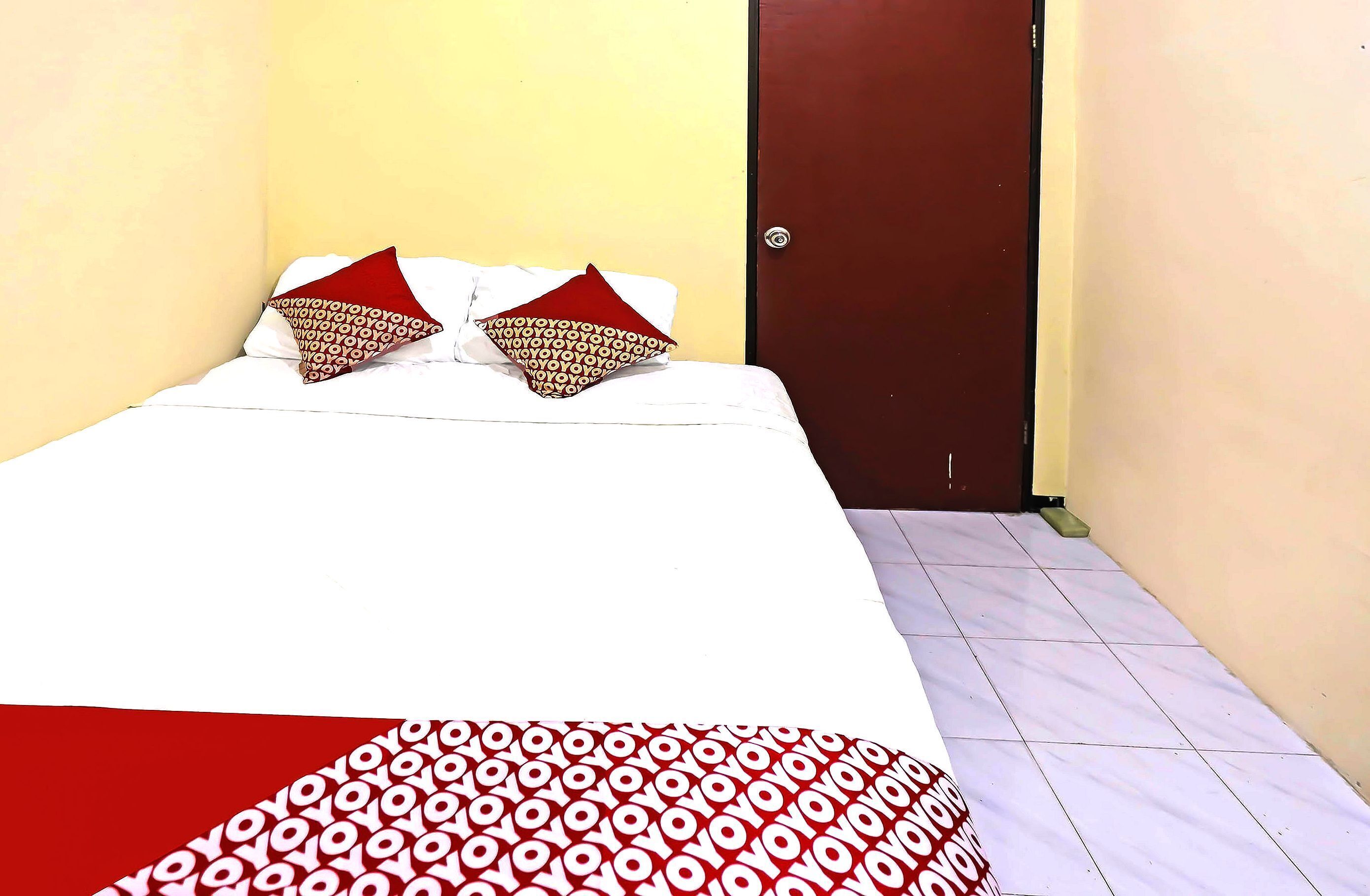 A small, neatly made bed with patterned pillows and a red and white bedspread in a room with yellow walls and a brown door.