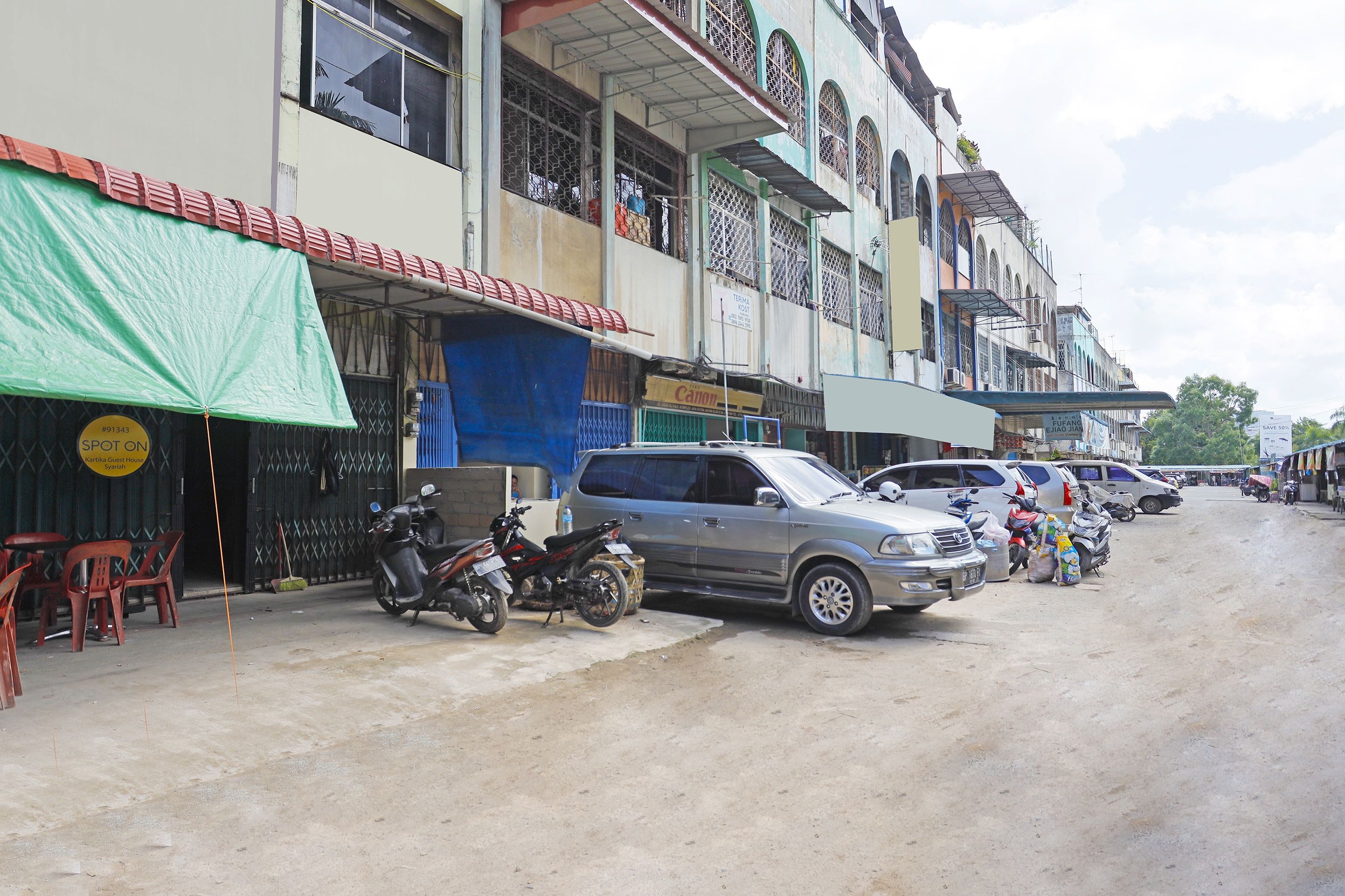 A street with parked cars and motorcycles, buildings with commercial signs, and a green tarp covering a structure.