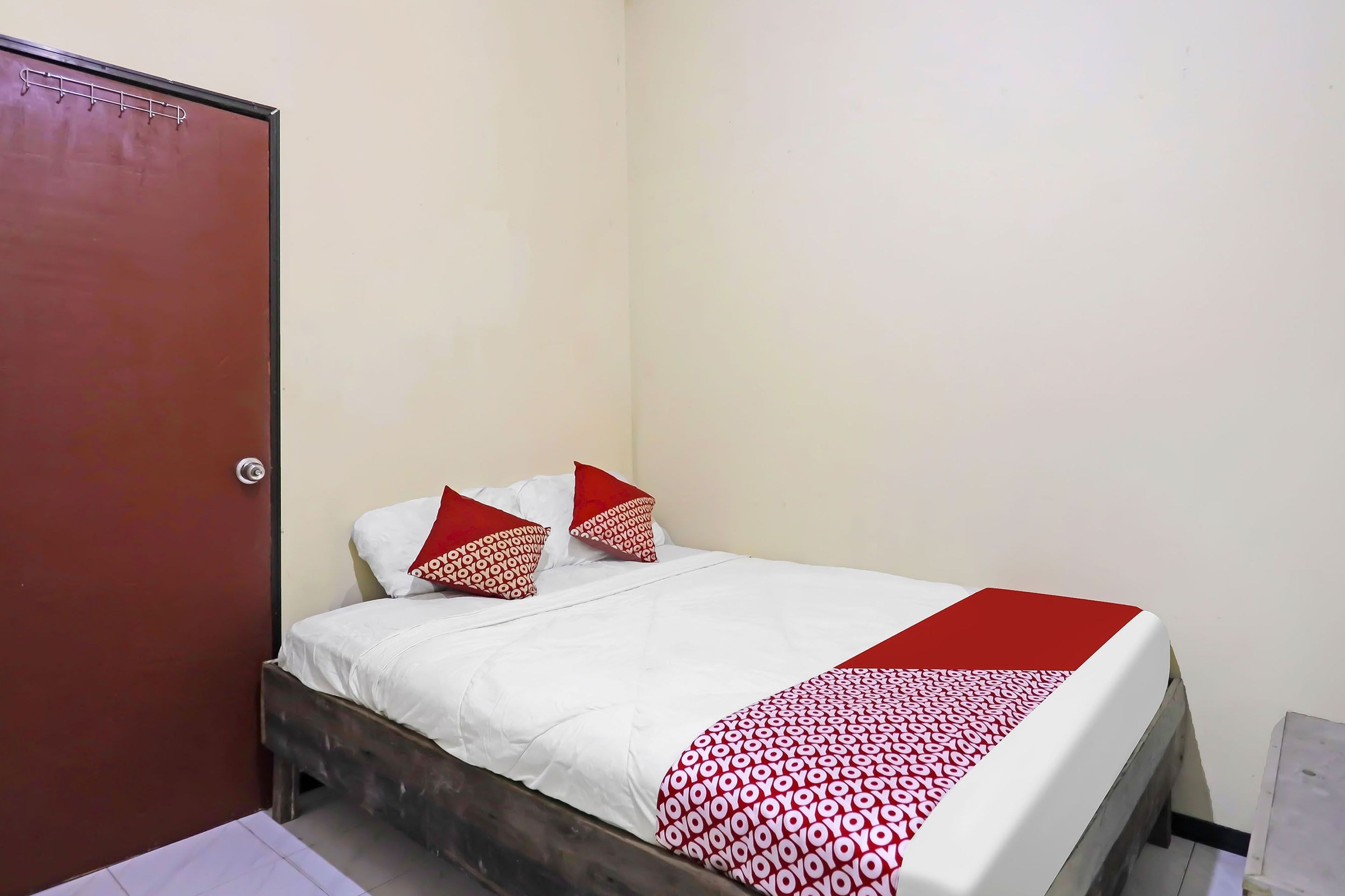 Uma cama de solteiro com roupa de cama branca e detalhes vermelhos, uma cabeceira de madeira e uma porta vermelha.