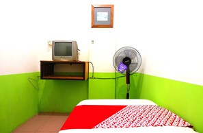A small room with a bed, a television on a shelf, and a standing fan. - Spot On 91343 Kartika Guest House Syariah (Batam)