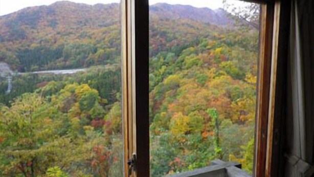 A room with a large window offering a view of a forested mountain range. - Pension Swiss (Kaminoyama)