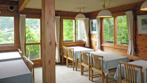 A dining area with wooden tables and chairs, large windows with curtains, and a view of greenery outside. - Pension Swiss (Kaminoyama)