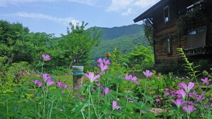 A rustic wooden cabin with a garden in front, surrounded by lush greenery and mountains in the background. - Pension Swiss (Kaminoyama)
