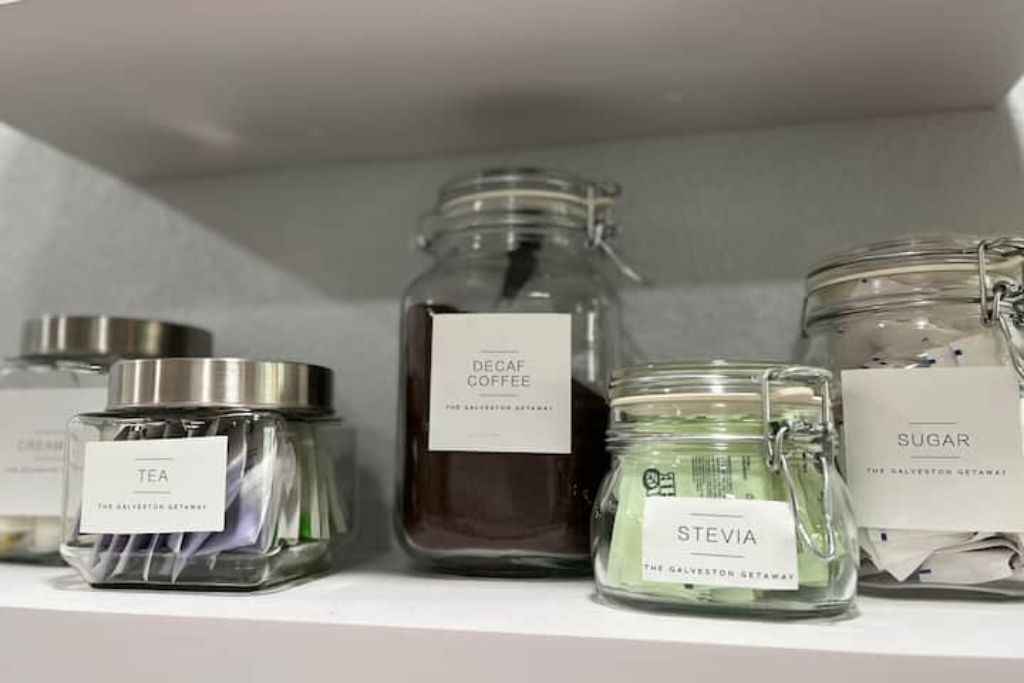 Una estantería con cuatro frascos de vidrio etiquetados como 'Tea', 'Decaf Coffee', 'Stevia' y 'Sugar'.