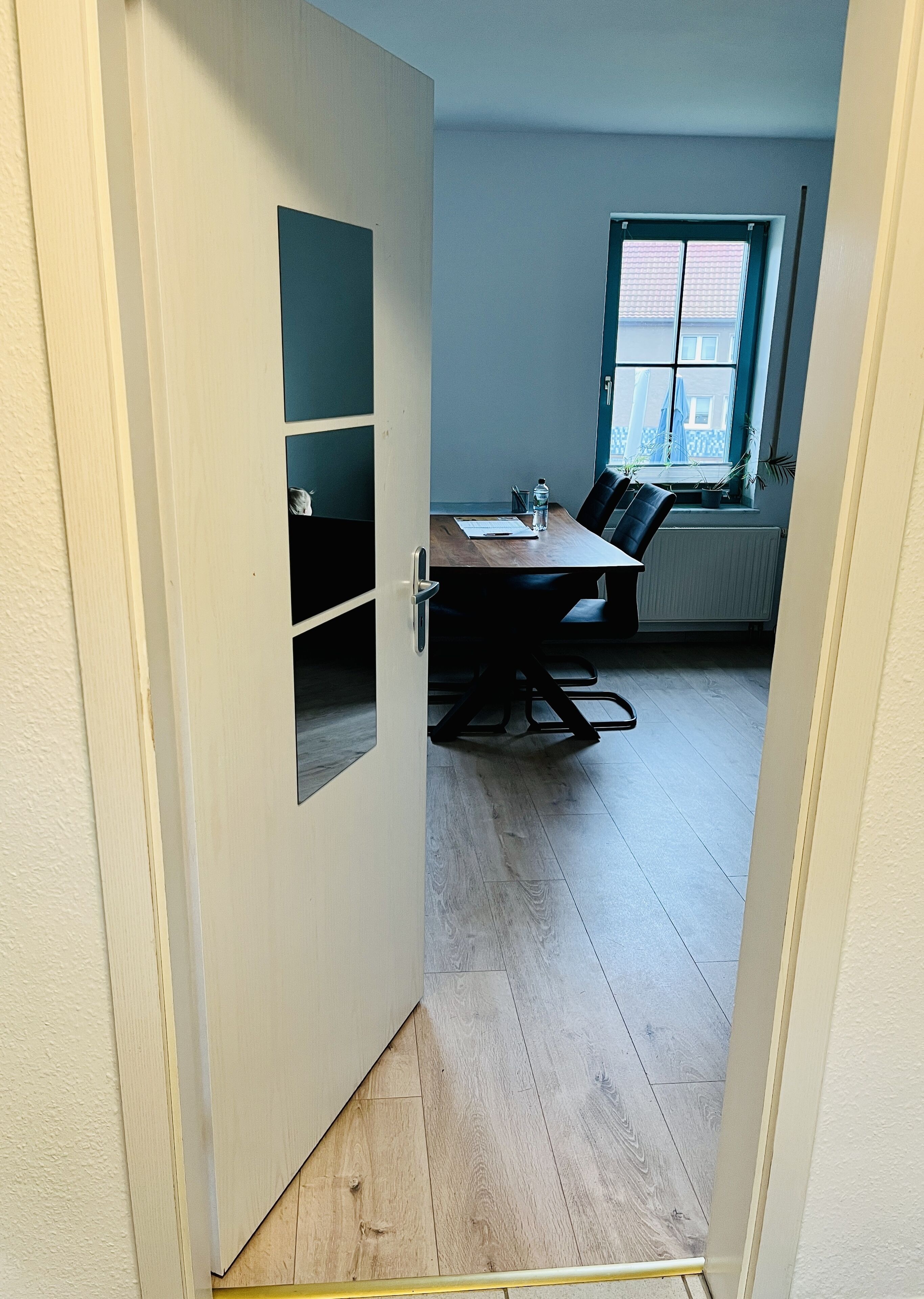 A room with a wooden floor, a table with chairs, a window with blinds, and a radiator.