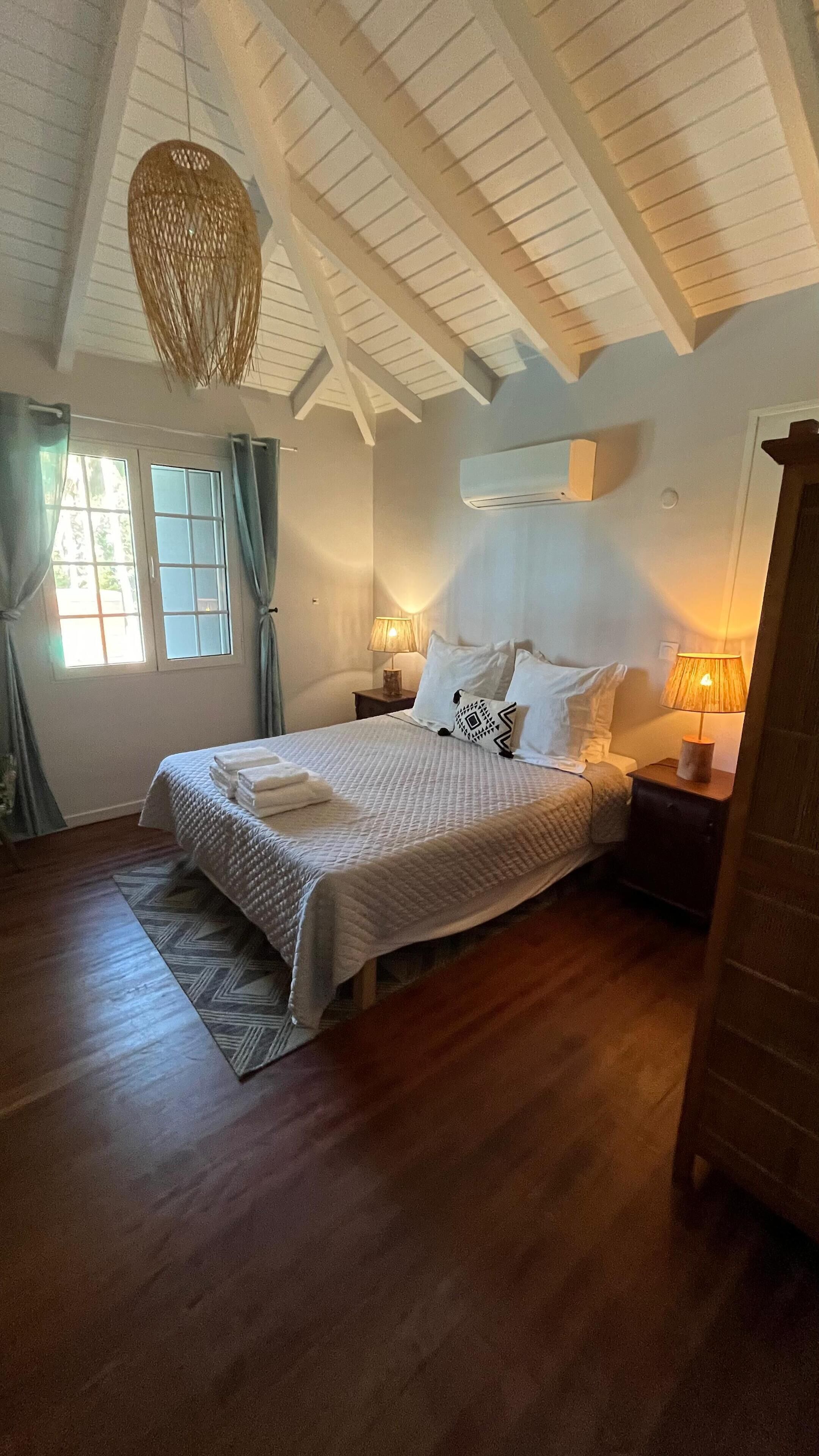 A bedroom with a large bed, a hanging light fixture, a window with curtains, and a wooden floor.