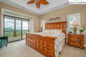 Un dormitorio con una cama grande, cabecera de madera, mesita de noche, lámpara y vistas a un balcón con barandilla de madera.