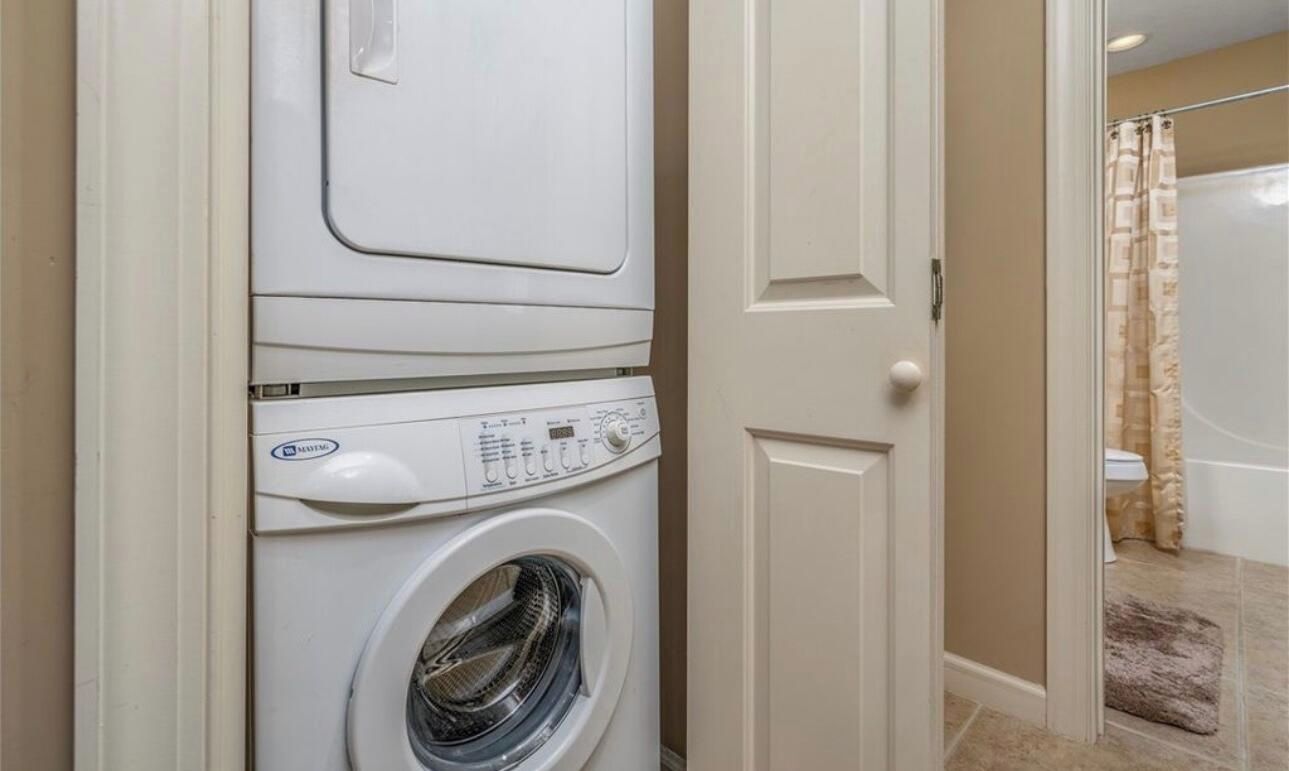 Een kleine wasruimte met een voorlader wasmachine en droger, een deur naar een badkamer met douche, en een klein vloerkleed.