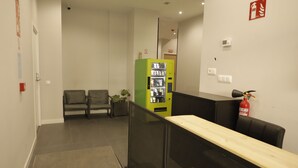 A reception area with a green vending machine, two grey sofas, a fire extinguisher, and a wooden reception desk. - MYD Madriz Hostel (Madrid)