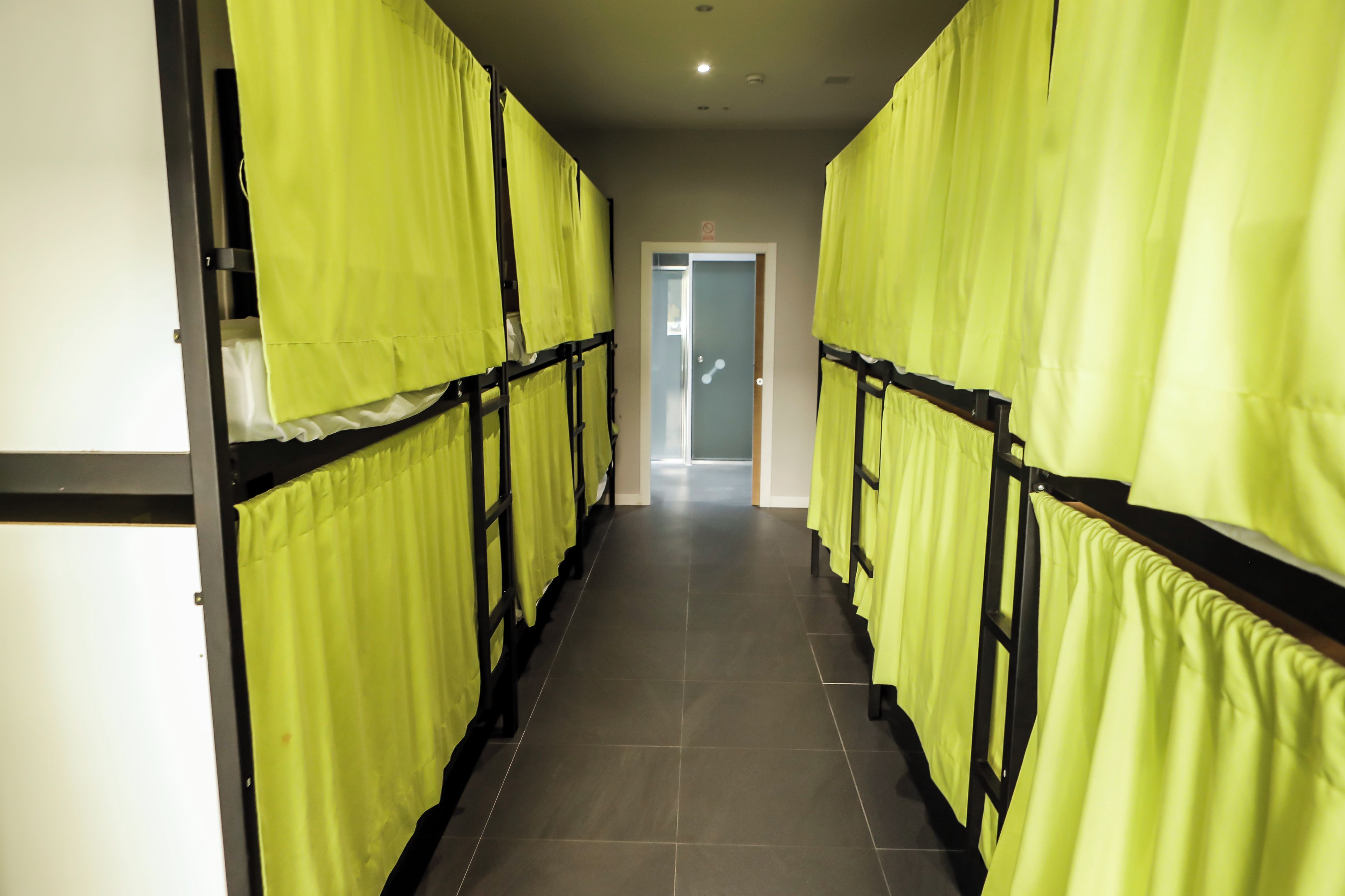 Un couloir bordé de lits superposés, chacun étant muni d’un rideau jaune, qui mène à une porte au fond.