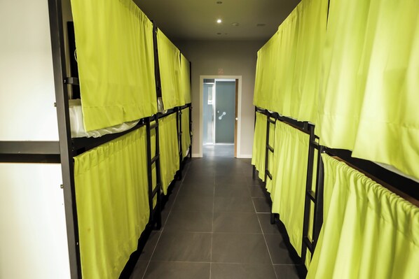 Un couloir bordé de lits superposés, chacun étant muni d’un rideau jaune, qui mène à une porte au fond.