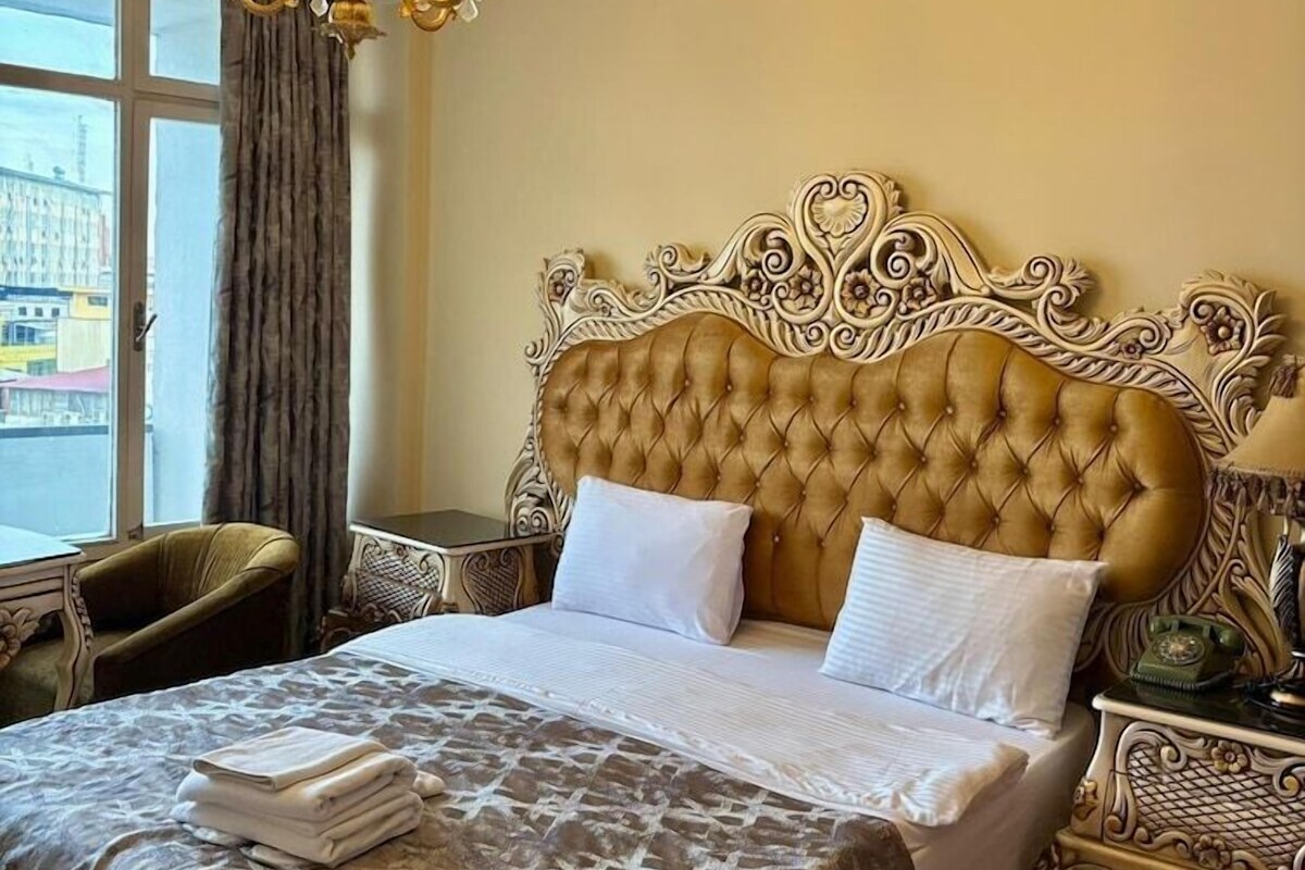 A hotel room with a tufted headboard, a chandelier, a bed with a patterned bedspread, a chair, and a nightstand with a lamp.