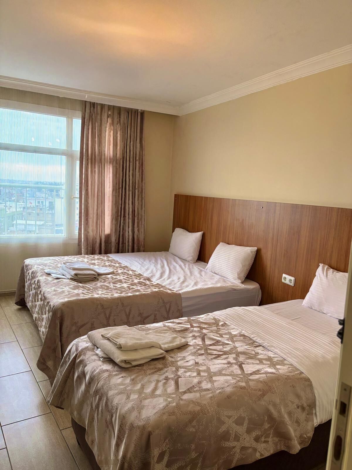 A hotel room with two beds, a large window with curtains, and a view of the city.