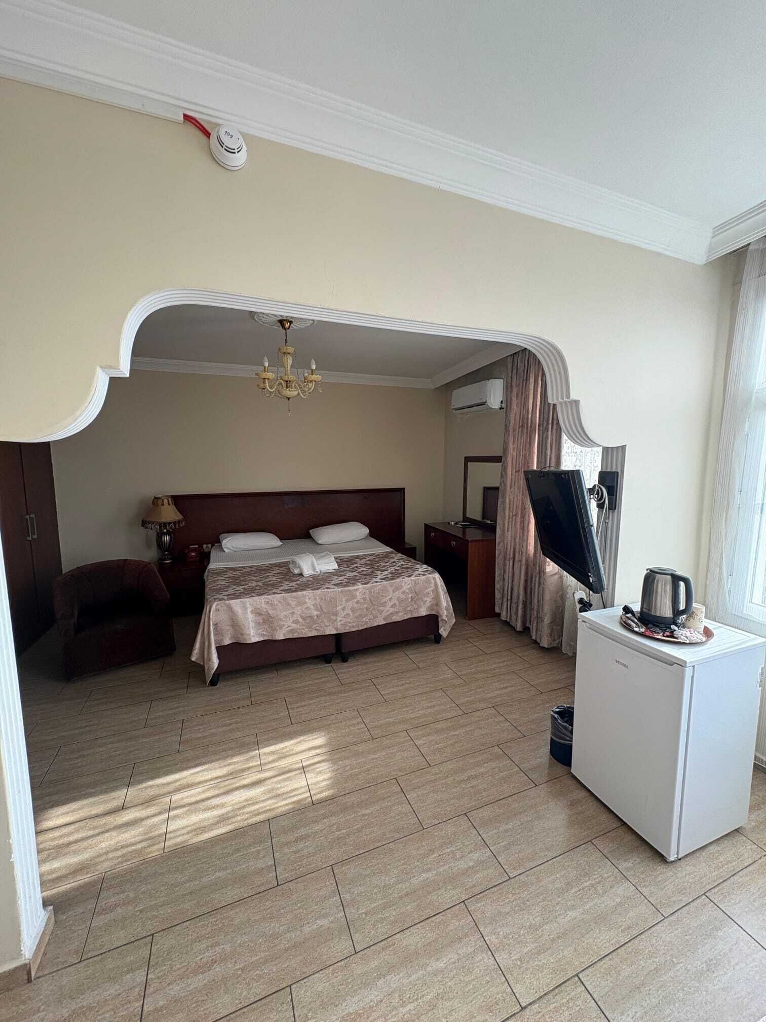 Habitación de hotel con cama, escritorio, silla, candelabro y un refrigerador pequeño.