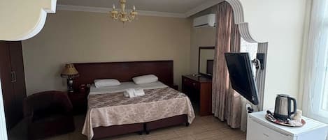 A hotel room with a bed, a desk, a chair, a chandelier, and a small refrigerator.
