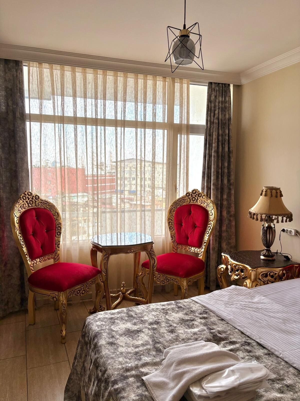 Habitación de hotel con dos sillones rojos tapizados, una mesita redonda pequeña, una cama con colcha gris estampada, una lámpara y vistas a edificios a través de cortinas de gasa.