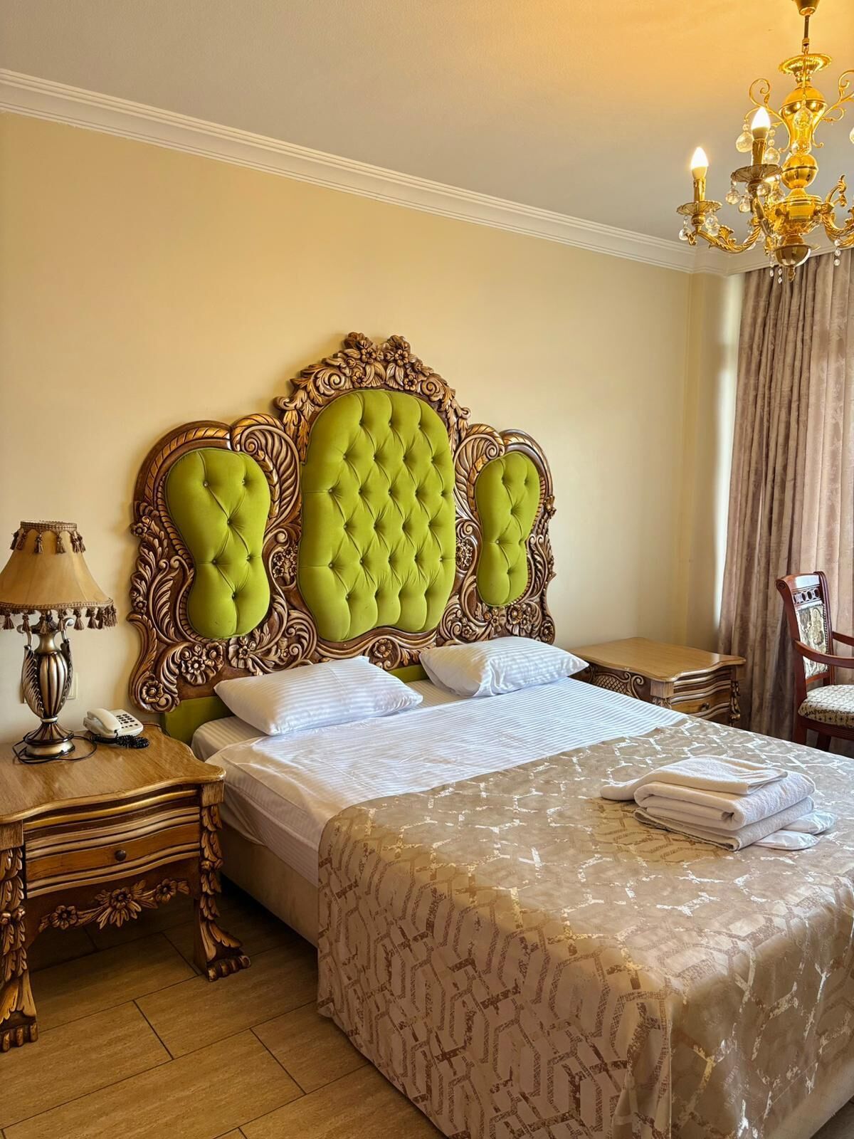 A hotel room with a large bed, ornate headboard, a chandelier, a bedside table with a lamp, and a chair.