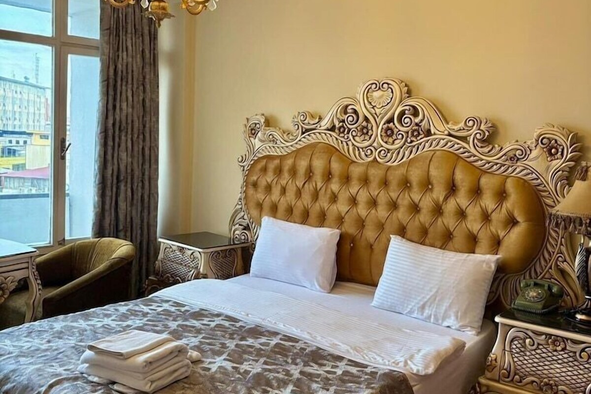 A hotel room with a tufted headboard, a chandelier, a bed with a patterned bedspread, a chair, and a nightstand with a lamp.