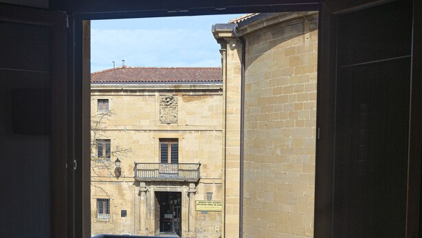 Vista a través de una puerta de madera con barandilla metálica, que da a un patio con suelo empedrado y un edificio con tejado de tejas al fondo.