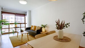 A modern living room with a sofa, coffee table, and dining table. There is a large window providing natural light and a view of the city.
