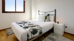 A neatly made bed with a white and green leaf-patterned bedspread, a wooden headboard, and a bedside table with a lamp.