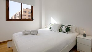 A modern bedroom with a large bed, white bedding, a nightstand with a lamp, and a window with a view of buildings. - Charming Madrid Airport III (Madrid)