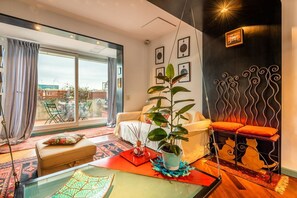 A modern living room with a large sliding glass door, a white sofa, a red ottoman, a glass coffee table, and a decorative metal wall piece.