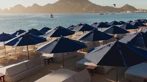 A beachside lounge area with multiple umbrellas and lounge chairs, overlooking a calm sea with boats and distant hills. - Corazon Cabo - Beachfront 2 bed/2 bath Suite with HOT TUB! (Cabo San Lucas)