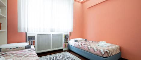 Un dormitorio con cama, mesita de noche, estantería, radiador y ventana con cortinas.