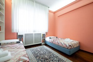 Un dormitorio con cama, mesita de noche, estantería, radiador y ventana con cortinas.