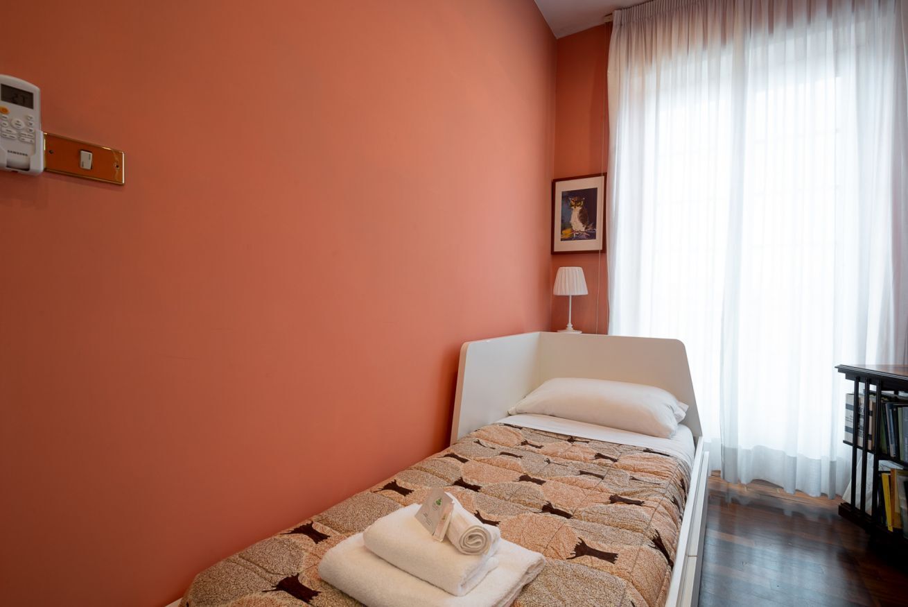 Una cama individual con colcha estampada, un termostato en la pared, un cuadro enmarcado y una ventana con cortinas de encaje.