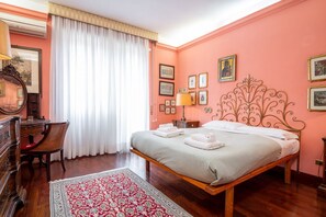 Ein Schlafzimmer mit einem hölzernen Bett, einem Schreibtisch mit Stuhl, einem großen Fenster mit Vorhängen und einem gemusterten Teppich.
