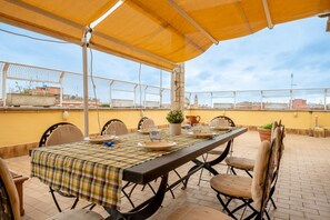 Eine Dachterrasse mit kariertem Tischleinentuch, Stühlen, einem Essplatz und einem gelben Sonnensegel.