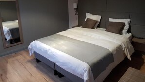 Una cama bien hecha con cabecera gris, una mesita de noche con lámpara y un espejo en la pared.