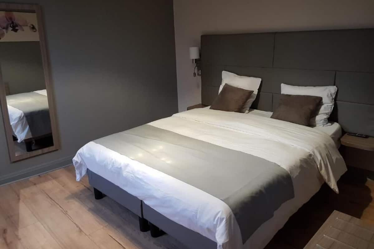 Una cama bien hecha con cabecera gris, una mesita de noche con lámpara y un espejo en la pared.