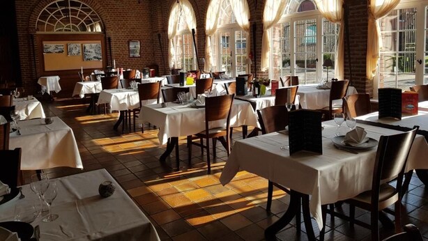 Un restaurante con varias mesas y sillas, paredes de ladrillo y amplios ventanales con cortinas.