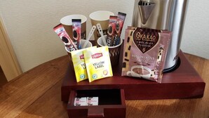 Una estación de café con cafetera, tazas, bolsitas de té Lipton y cacao en polvo Okakao.
