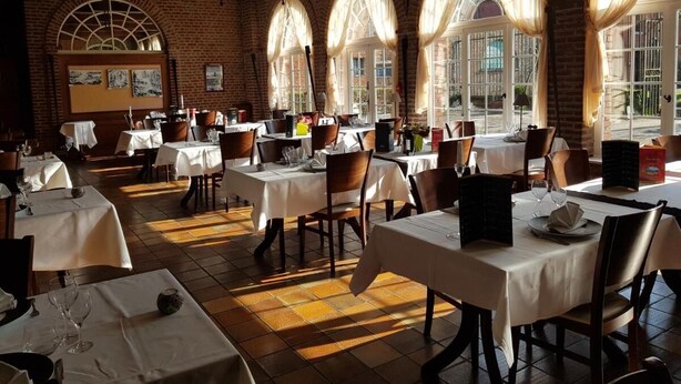 Un restaurante con varias mesas y sillas, paredes de ladrillo y amplios ventanales con cortinas.