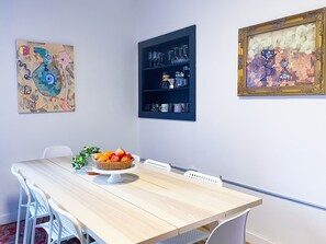 A dining area with a wooden table, white chairs, a fruit basket, and wall-mounted shelves with glassware and kitchen items. - The Little Black House - in the heart of RiNo! (Denver)