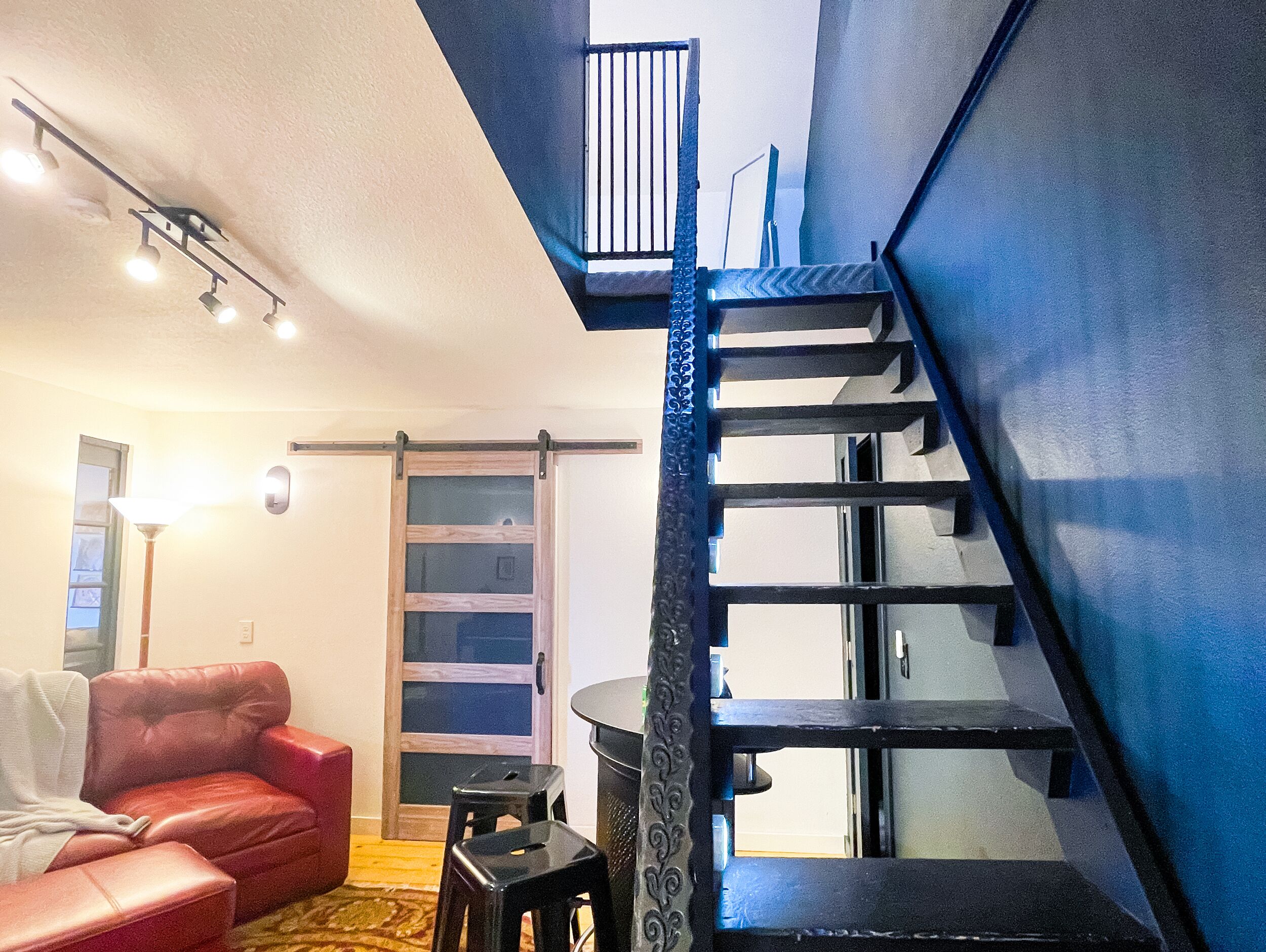 Una sala de estar moderna con un sofá de cuero rojo, un taburete negro pequeño y una escalera con barandilla azul que sube a un nivel superior.