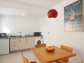 Una cocina compacta equipada con microondas, tostadora y pileta. Se observa una mesa de madera con un bol de fruta y dos sillas. Una lámpara colgante roja ilumina la mesa.