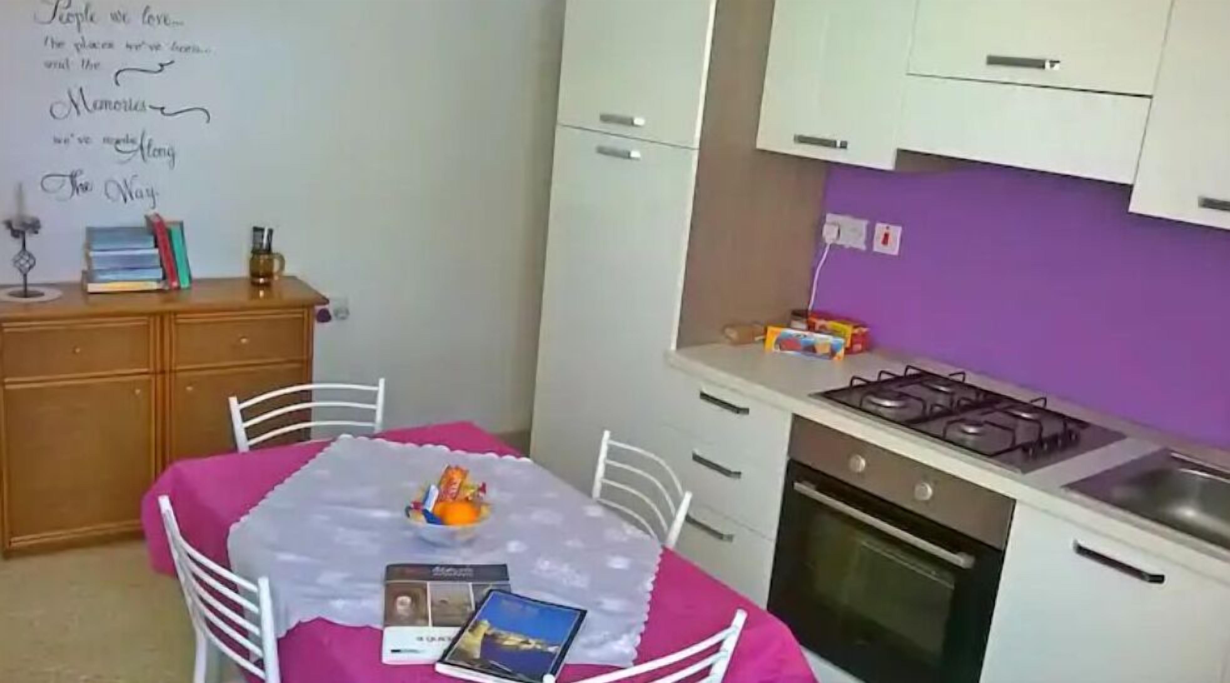 Une cuisine compacte équipée d’une cuisinière à gaz, d’un four et de meubles blancs. On y trouve une table à manger avec des chaises, ainsi qu’un mur violet orné d’un mot écrit à la main.